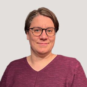 Das Portrait zeigt Christina A. Benker. Sie hat kurze braune Haare und trägt eine Brille. Sie lächelt in die Kamera.