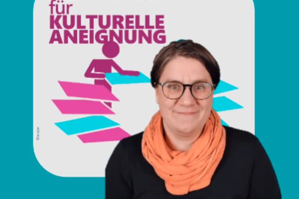 Eine Frau mit kurzen dunkelblonden Haaren vor einem dunkelpetrolfarbenen Hintergrund. Sie trägt ein schwarzes Oberteil mit einem orangefarbenen Schal. Sie hat eine Brille.